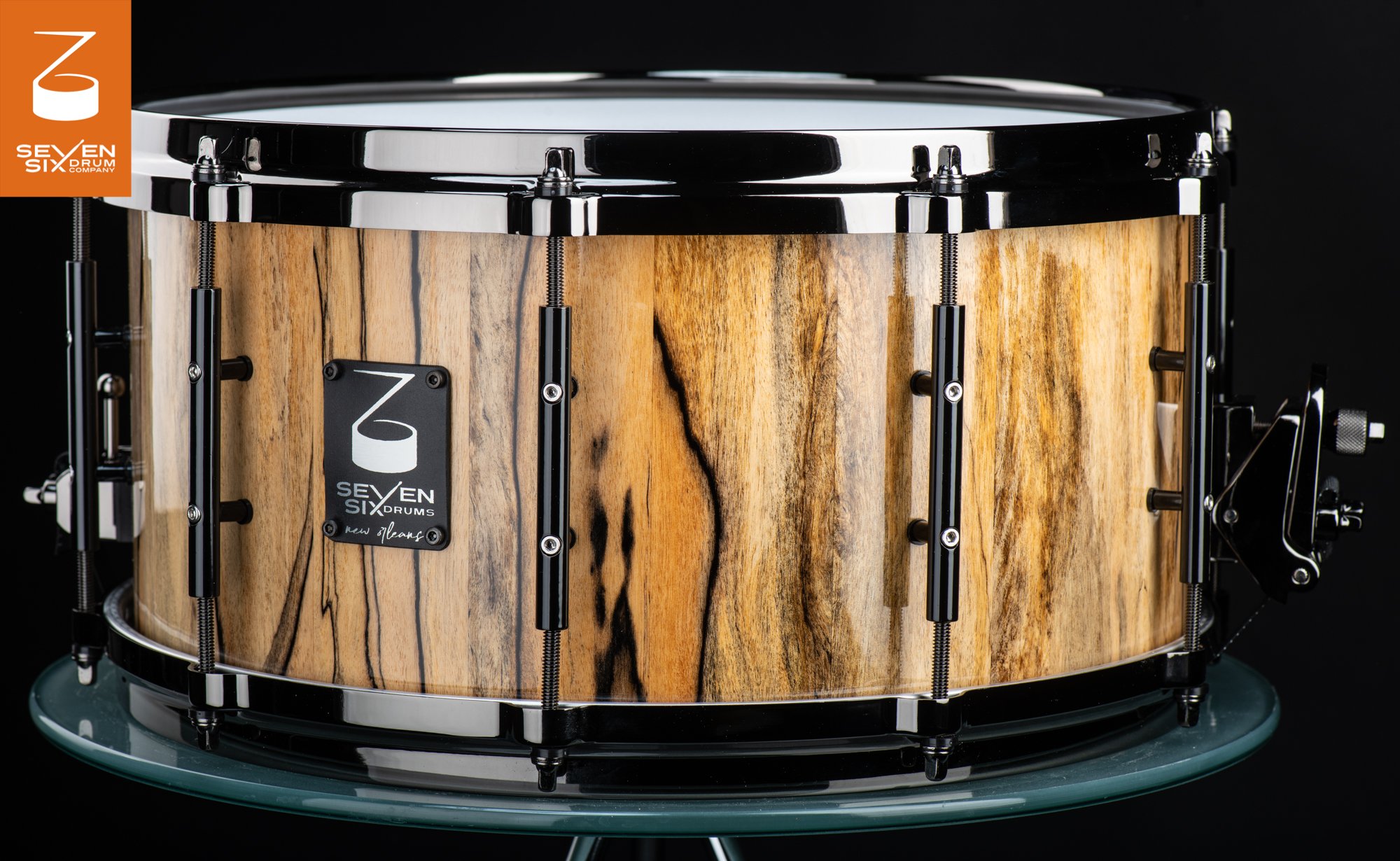 Handmade snare drum with a glossy, dark-grained natural wood shell, black nickel hoops, and black tube lugs.