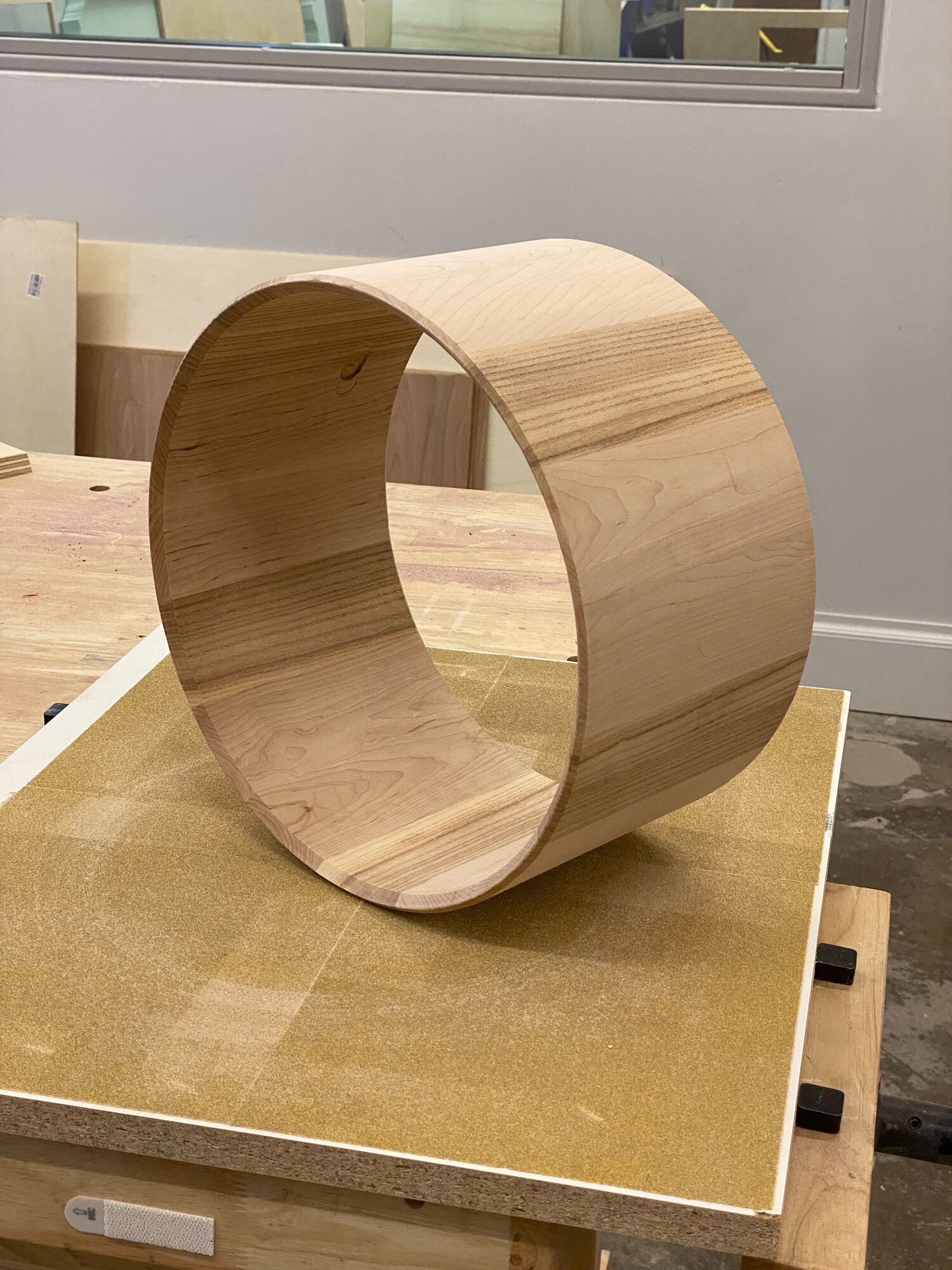 An unfinished natural light wood snare drum shell with no hardware rests upright on a workshop sanding board.