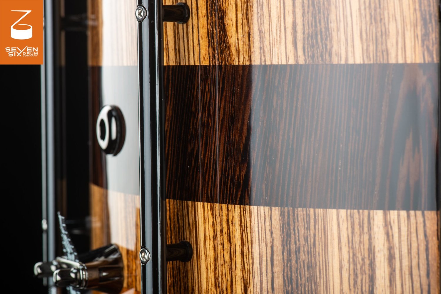 Close-up of a handcrafted snare drum with a high-gloss striped wood shell, black tube lugs, and a chrome throw-off.