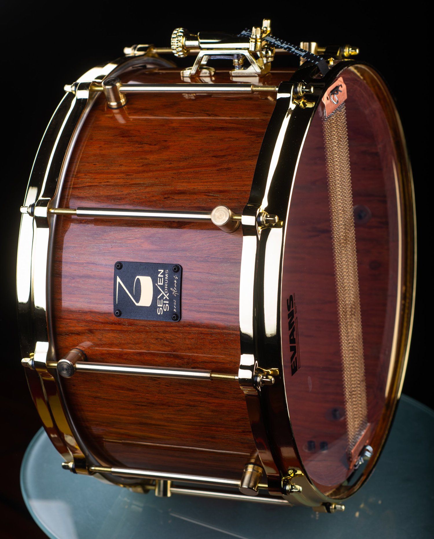 Deep snare drum with a glossy reddish-brown wood shell, polished gold hoops, tube lugs, and brass snare wires.