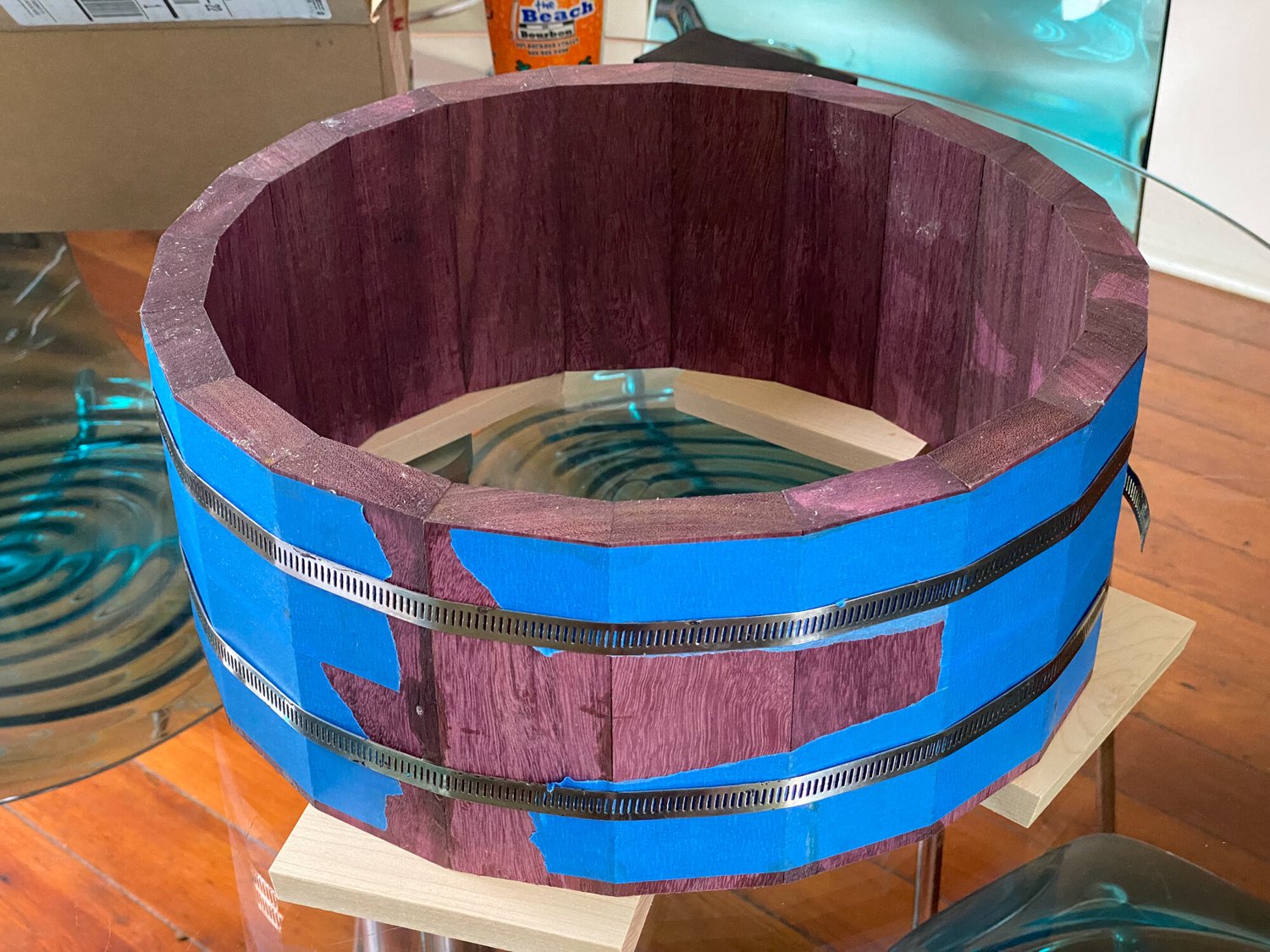 An unfinished purpleheart stave snare drum shell secured with blue painter's tape and two metal hose clamps.
