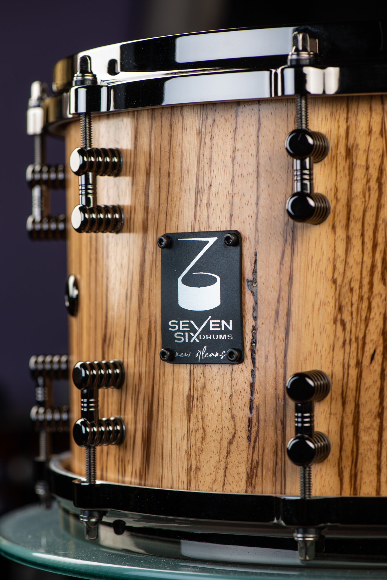 Handcrafted snare drum with a natural wood grain finish, glossy black nickel hoops, ribbed lugs, and a black maker's badge.