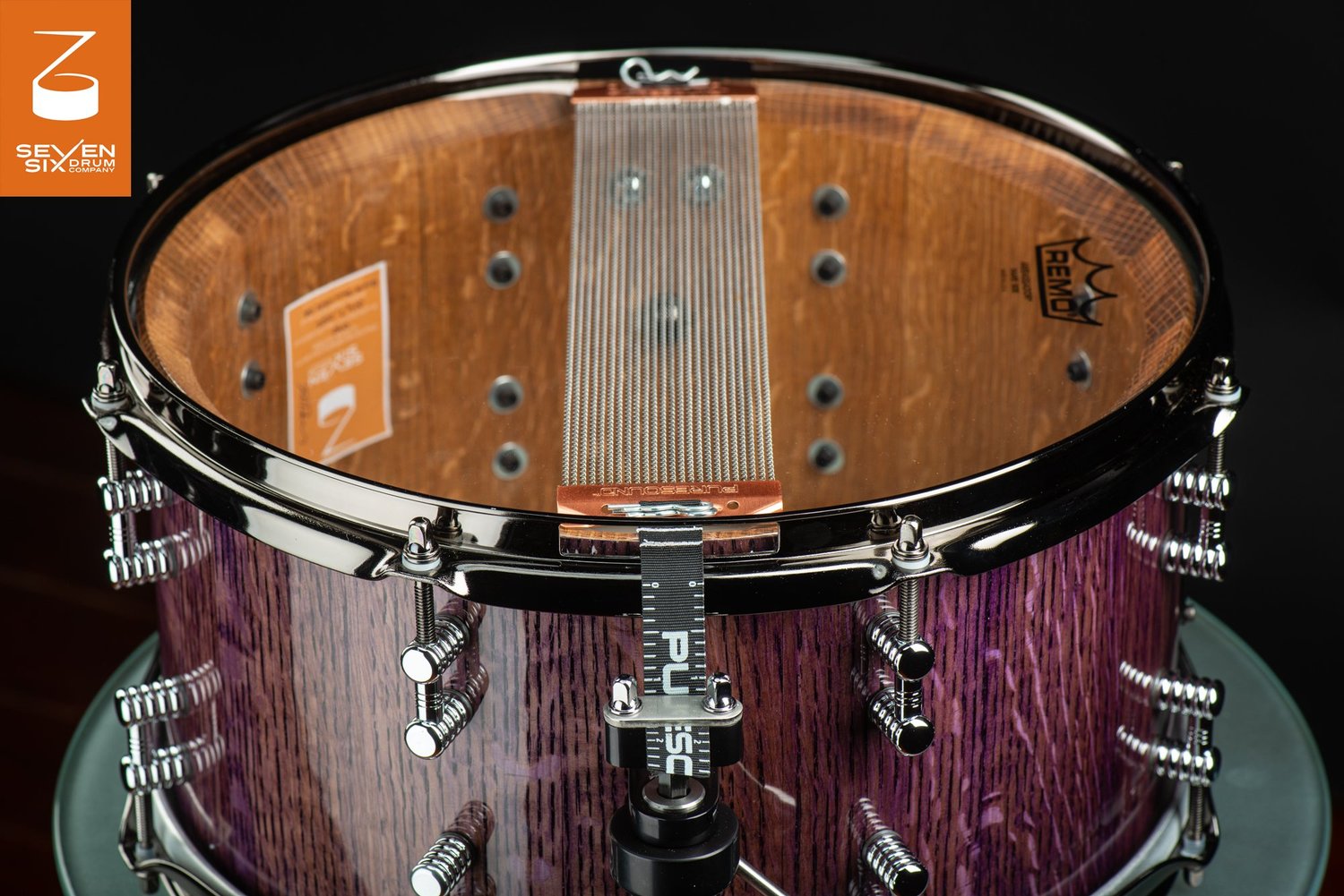 Glossy purple-stained wood snare drum with chrome hardware, dual-ended lugs, and snare wires visible over a clear drumhead.