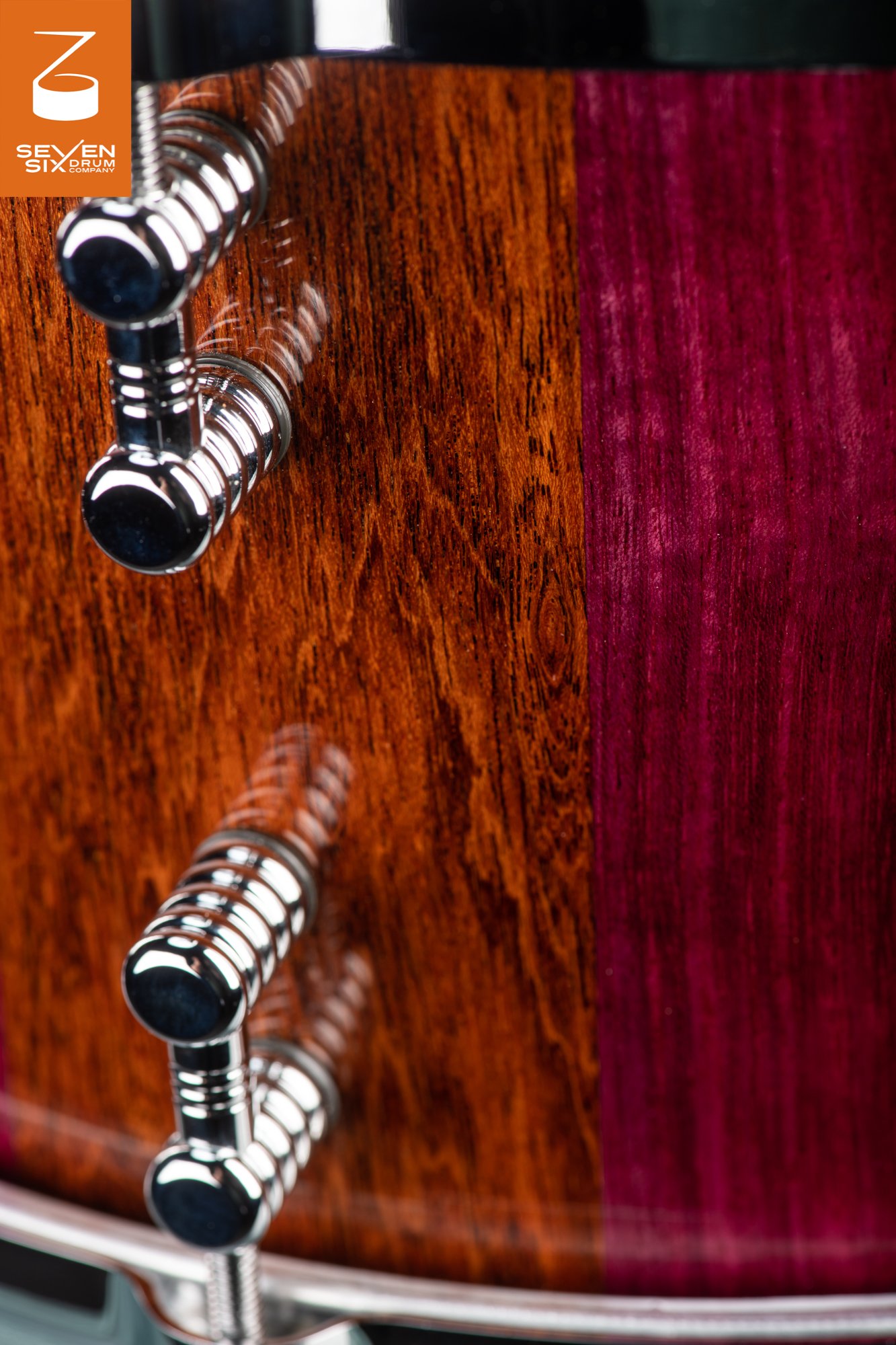 Close-up of a snare drum shell in glossy brown and purple wood with polished chrome tube lugs.