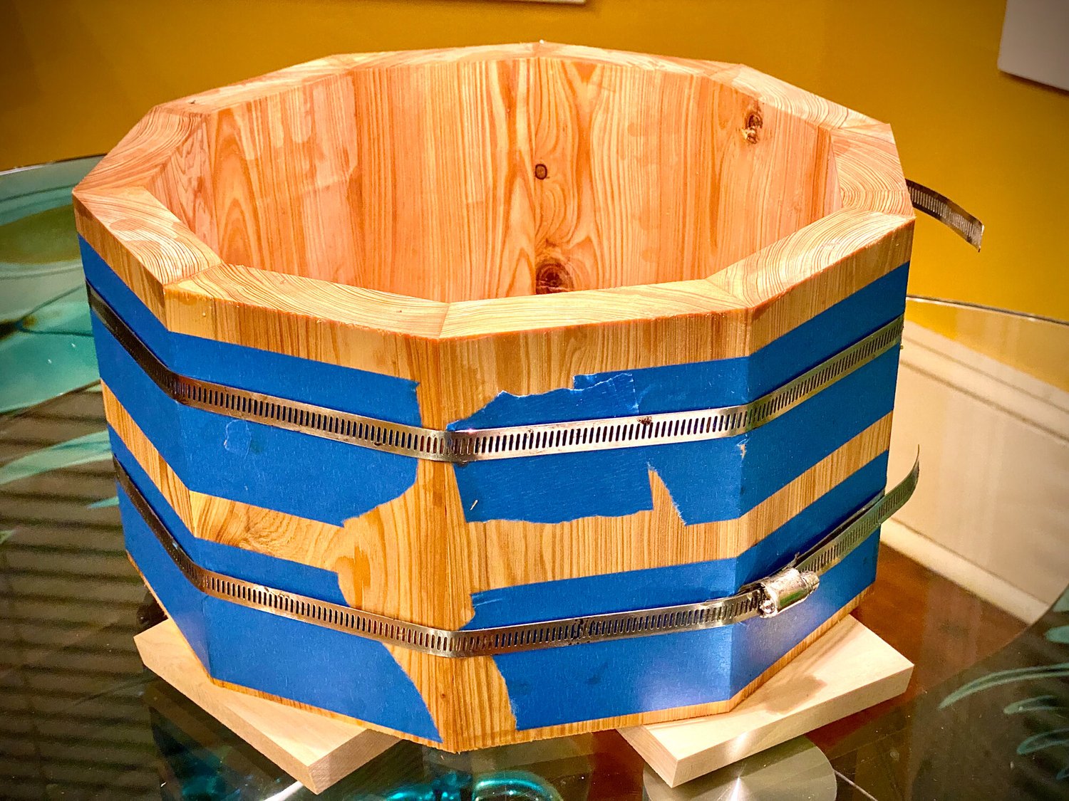 Unfinished natural pine stave drum shell clamped during assembly with blue painter's tape and metal hose clamps.