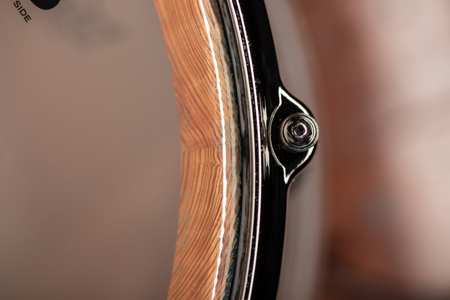 Detail of a snare drum with a natural wood shell interior, drumhead, and glossy black hoop with a chrome tension rod.