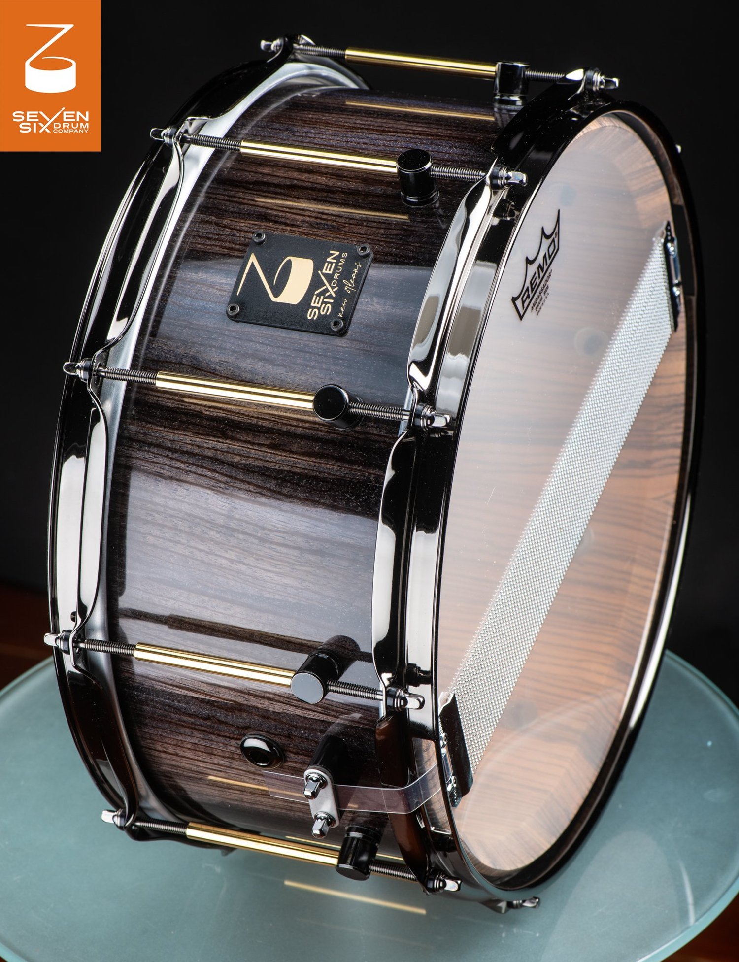 Handmade snare drum featuring a glossy dark wood grain shell, black chrome hoops, and brass tube lugs.