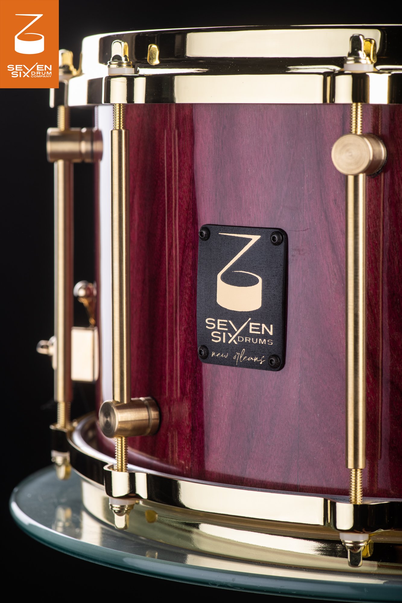 Handmade snare drum featuring a glossy purpleheart wood shell, brass tube lugs, matching brass hoops, and a black logo badge.
