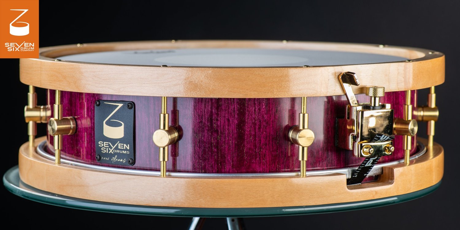 Handmade snare drum with a glossy purpleheart wood shell, natural maple wood hoops, brass lugs, and a gold throw-off.