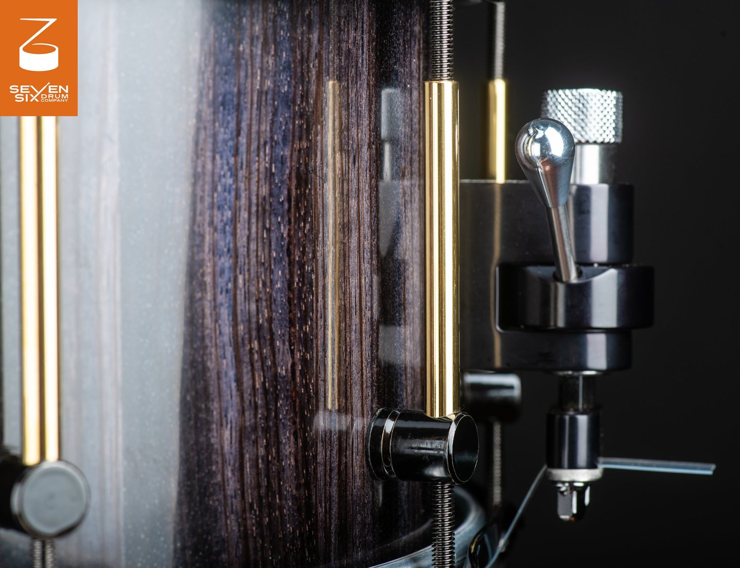 Close-up of a snare drum with a glossy dark wood shell, brass tube lugs, and a black and chrome throw-off.