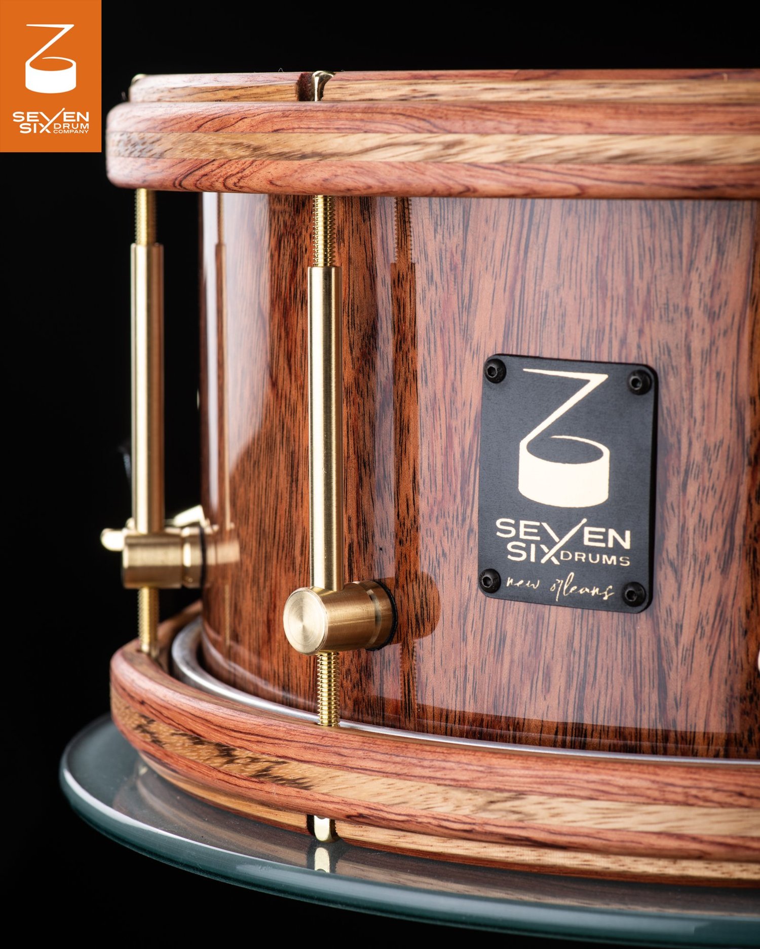 Glossy wood snare drum featuring striped wooden hoops, brass tube lugs, and a black metal logo badge.