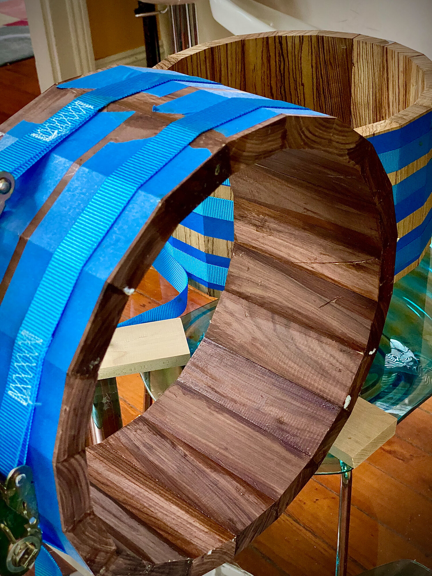 Unfinished walnut stave drum shell secured by blue ratchet strap clamps with metal buckles.