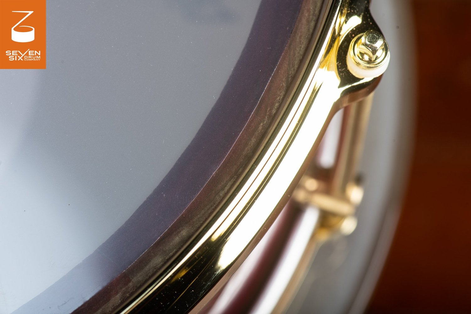 Close-up of a dark wood snare drum with a polished gold hoop, matching tension rod, and a clear drumhead.