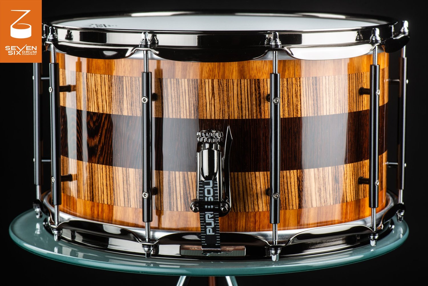 Glossy segmented wood snare drum with horizontal bands of grain, fitted with black nickel tube lugs and a throw-off.