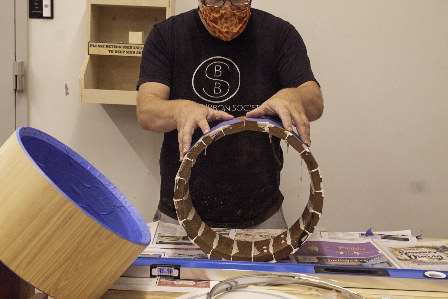 A drum builder glues an unfinished wooden stave shell at a workbench with a chrome drum hoop in the foreground.
