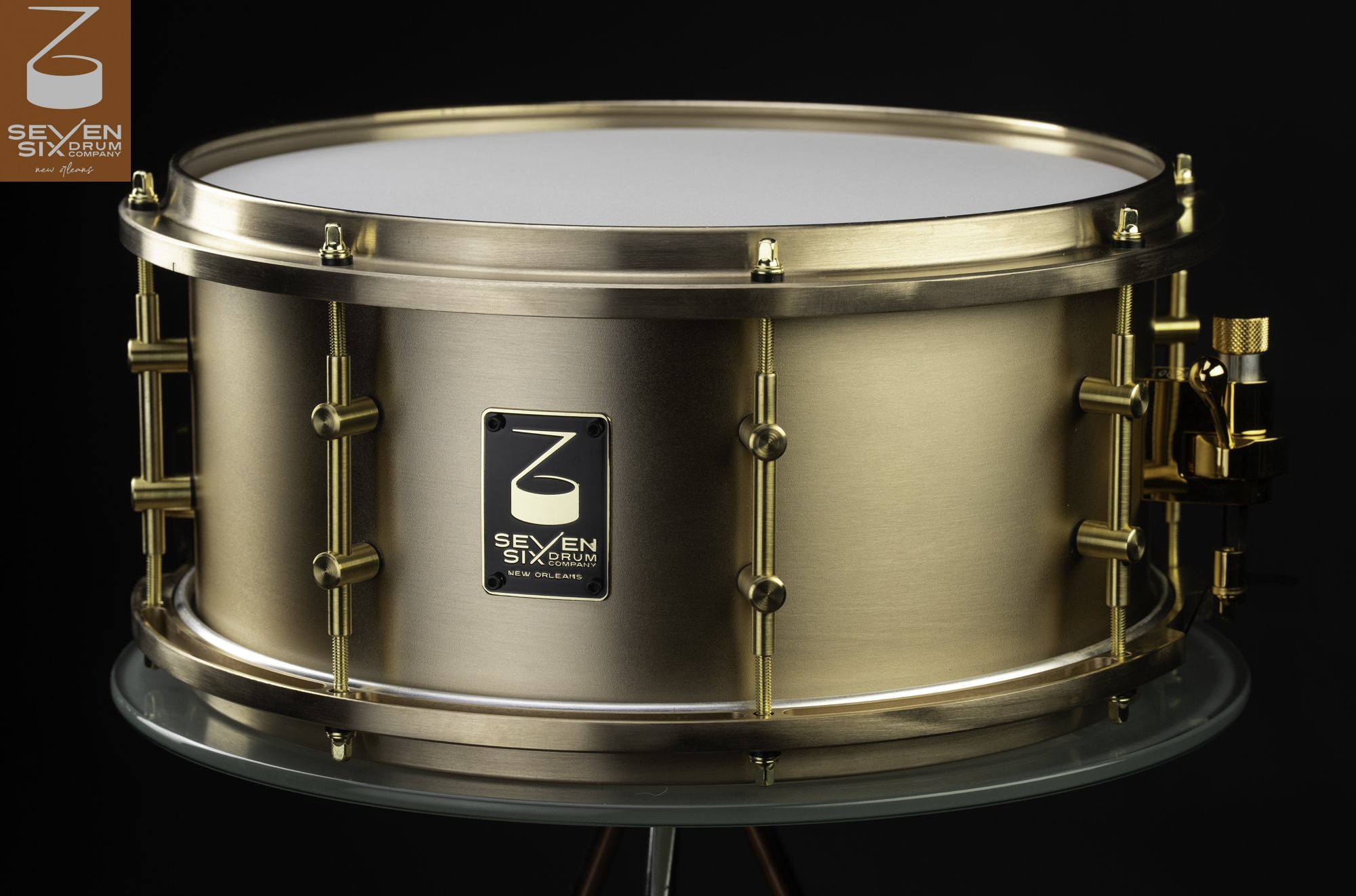 Brushed bronze snare drum with matching metal hoops, brass tube lugs, and a gold throw-off.