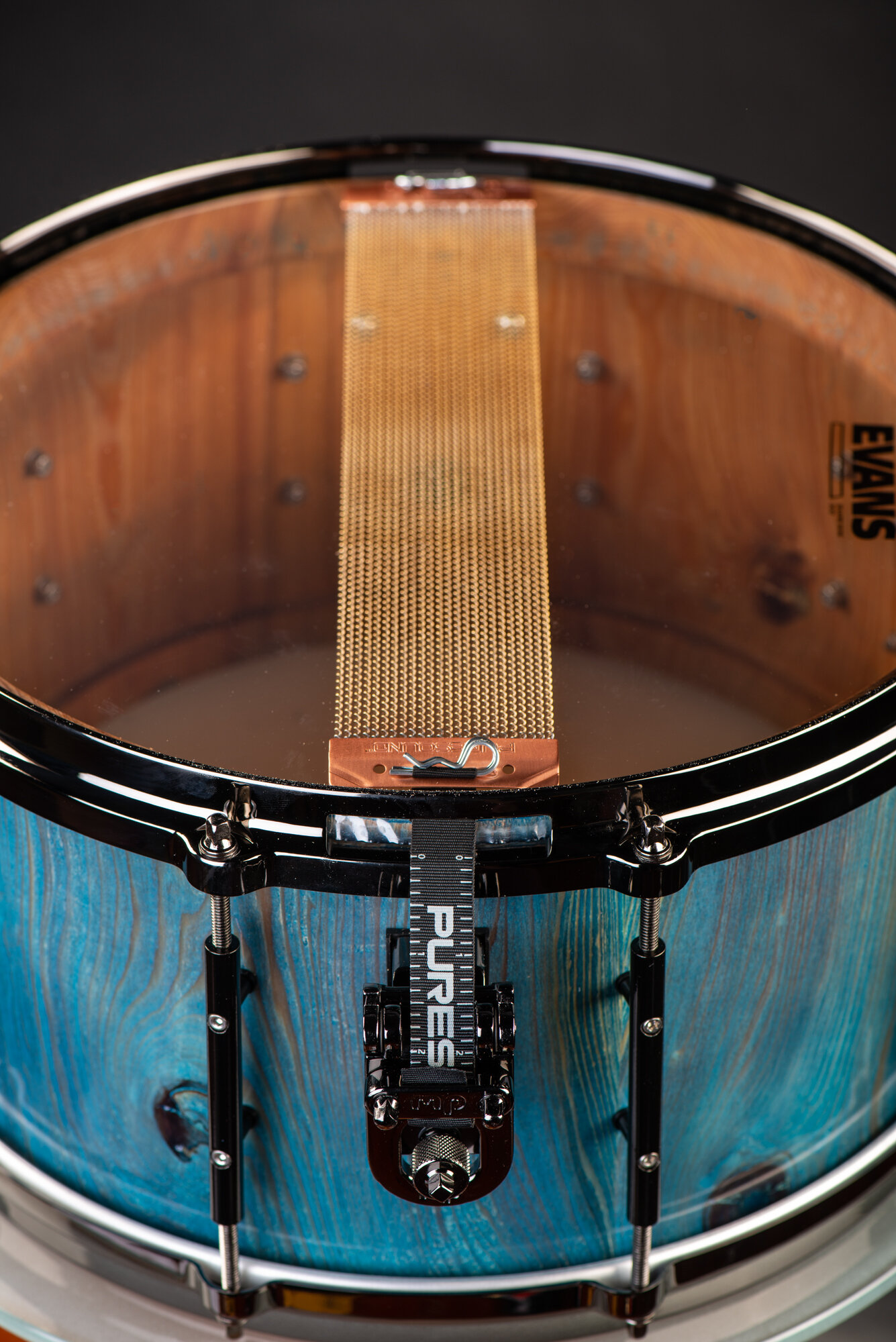 Underside of a blue-stained wood snare drum with black hardware, a DW throw-off, and wide brass snare wires.