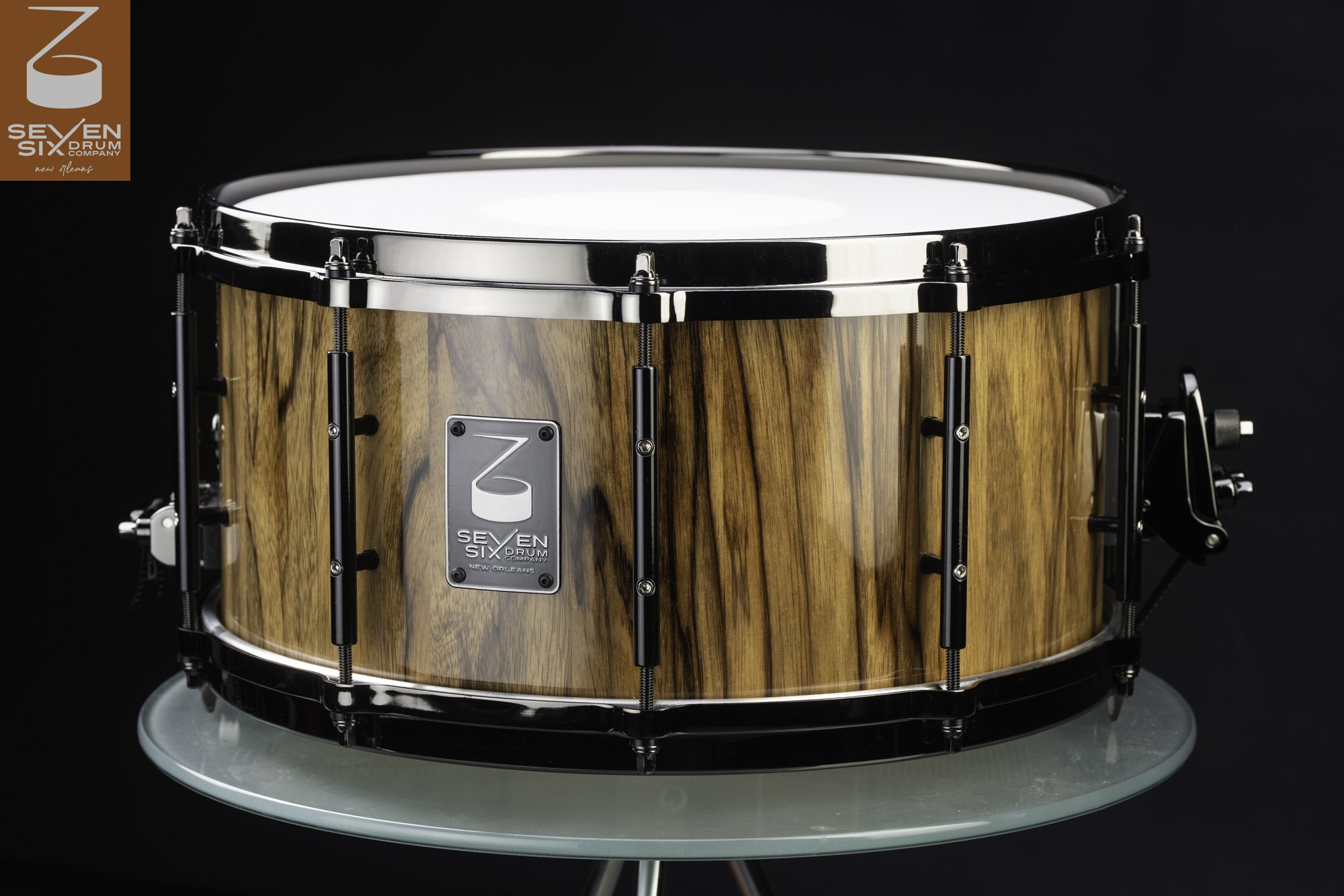 A glossy wood snare drum featuring prominent dark grain, polished chrome hoops, and black tube lugs.