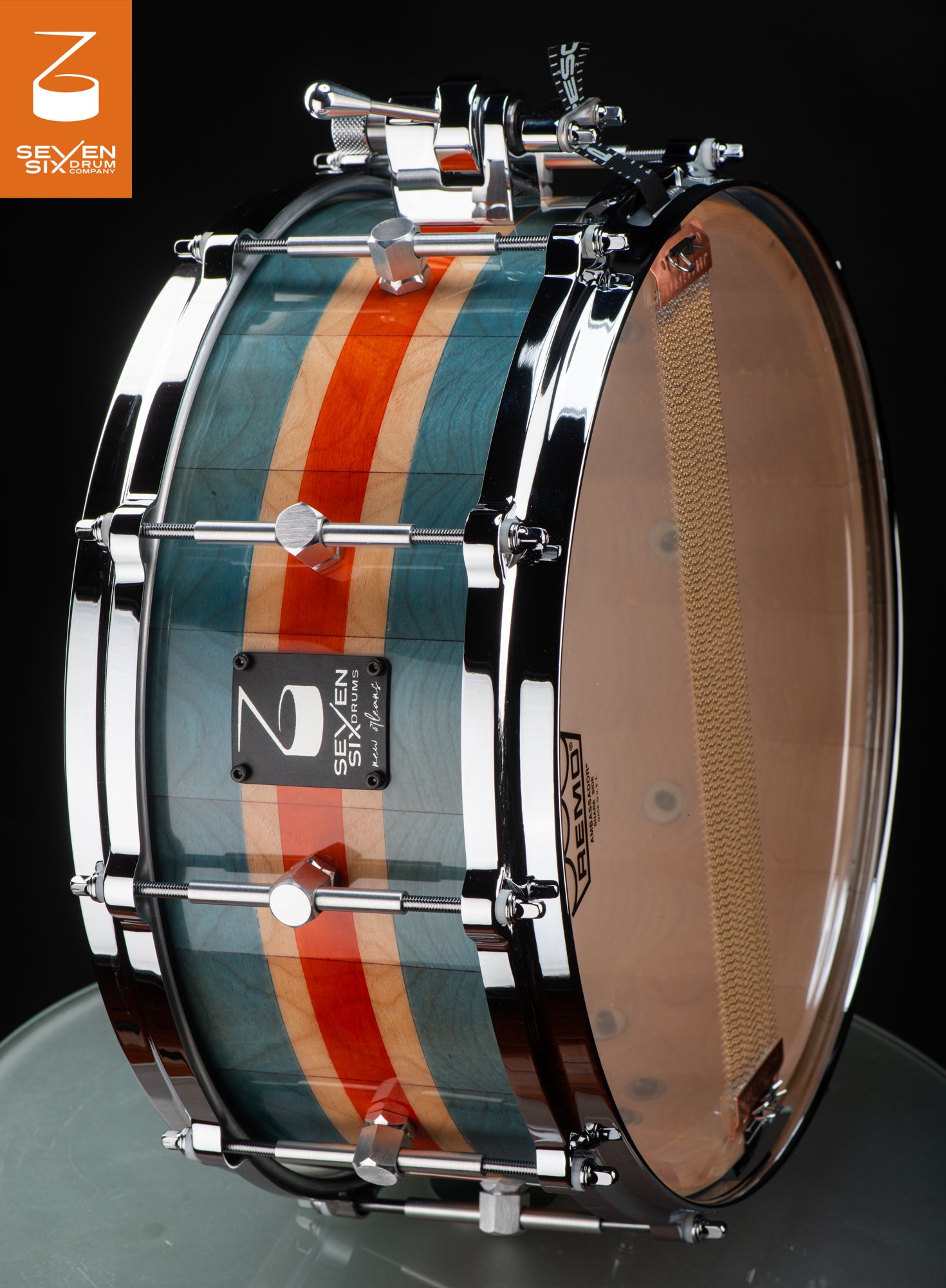 Handmade snare drum with a teal and orange striped wood shell, chrome hoops, center-mounted lugs, and brass snare wires.