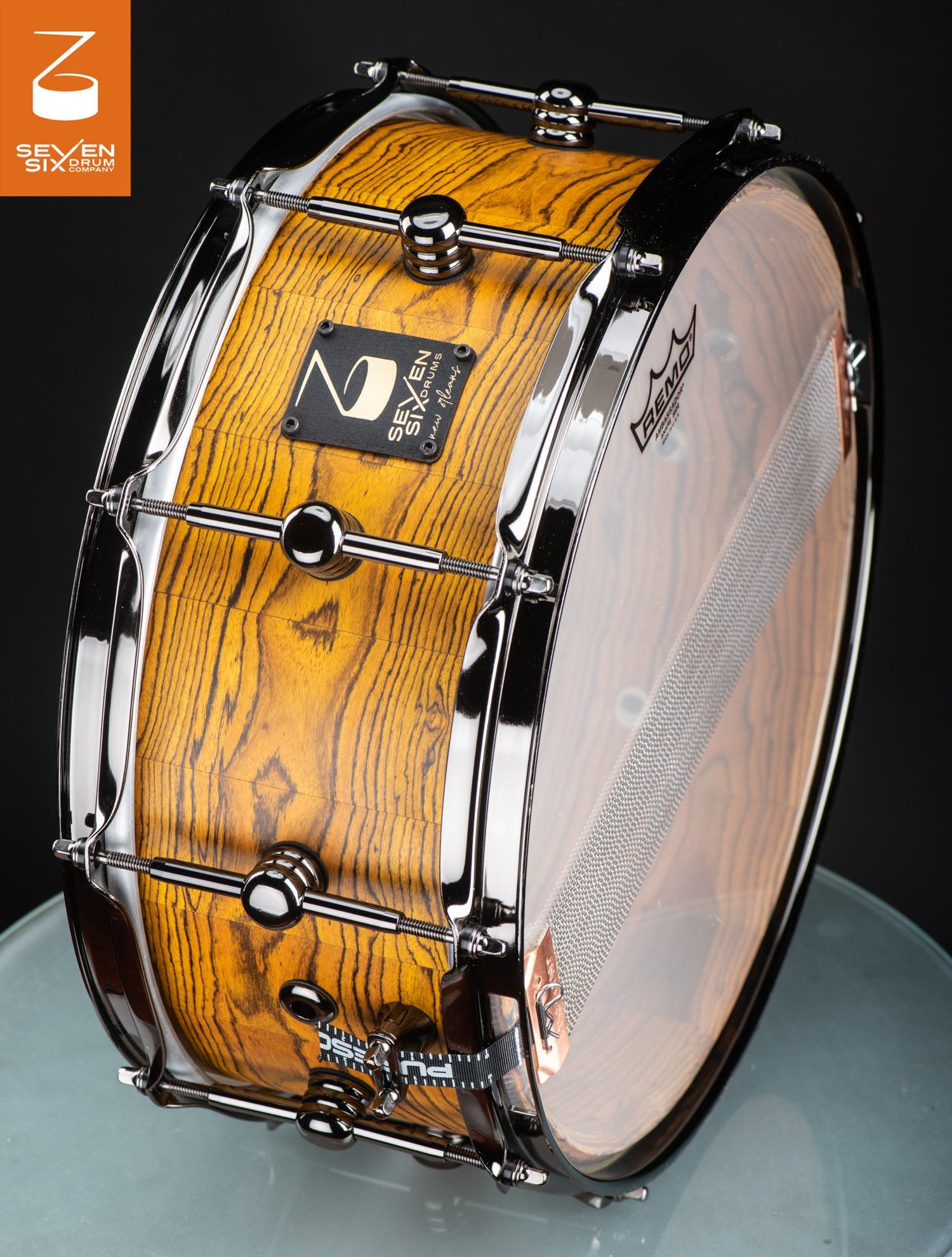 Handmade snare drum with a natural yellow wood grain shell, polished black chrome hoops, and center-mounted tube lugs.