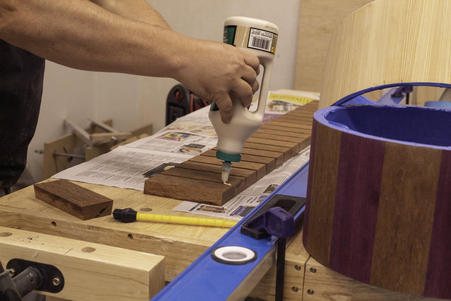 A craftsman applies wood glue to unfinished hardwood staves beside a clamped purpleheart snare drum shell.