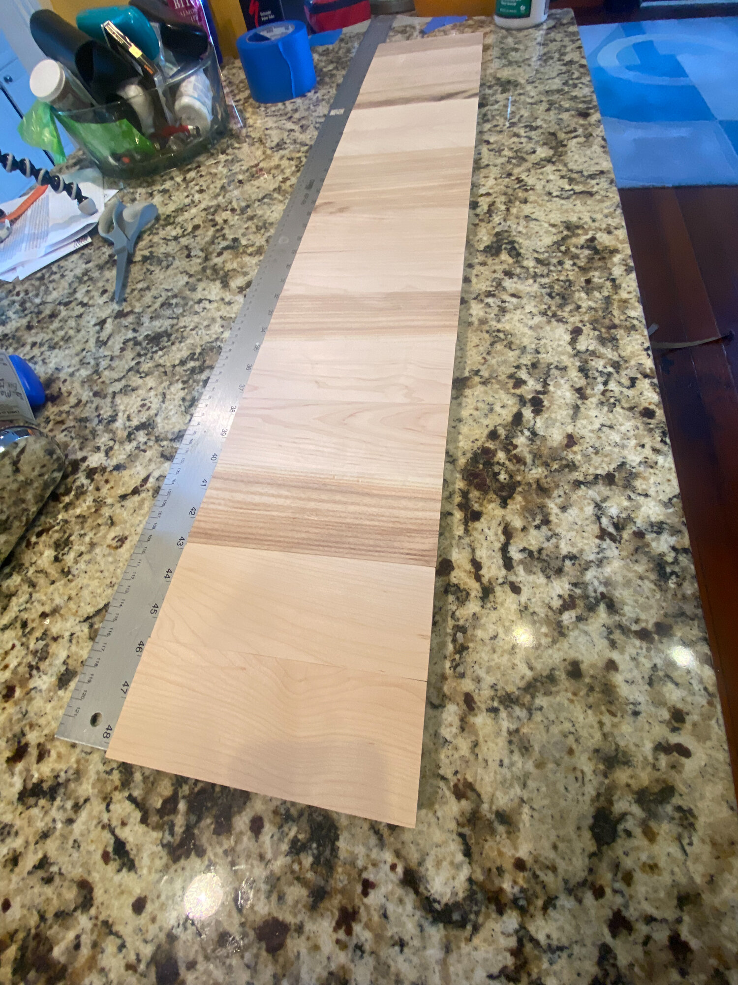 Unfinished light wood staves arranged beside a long metal ruler on a granite countertop.