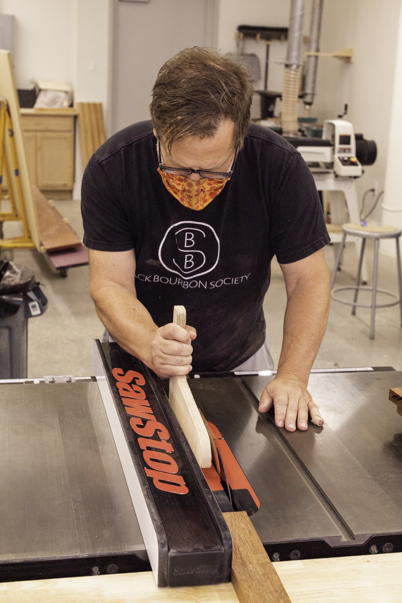 A craftsman cuts an unfinished hardwood board using a wooden push stick on a SawStop table saw in his workshop.