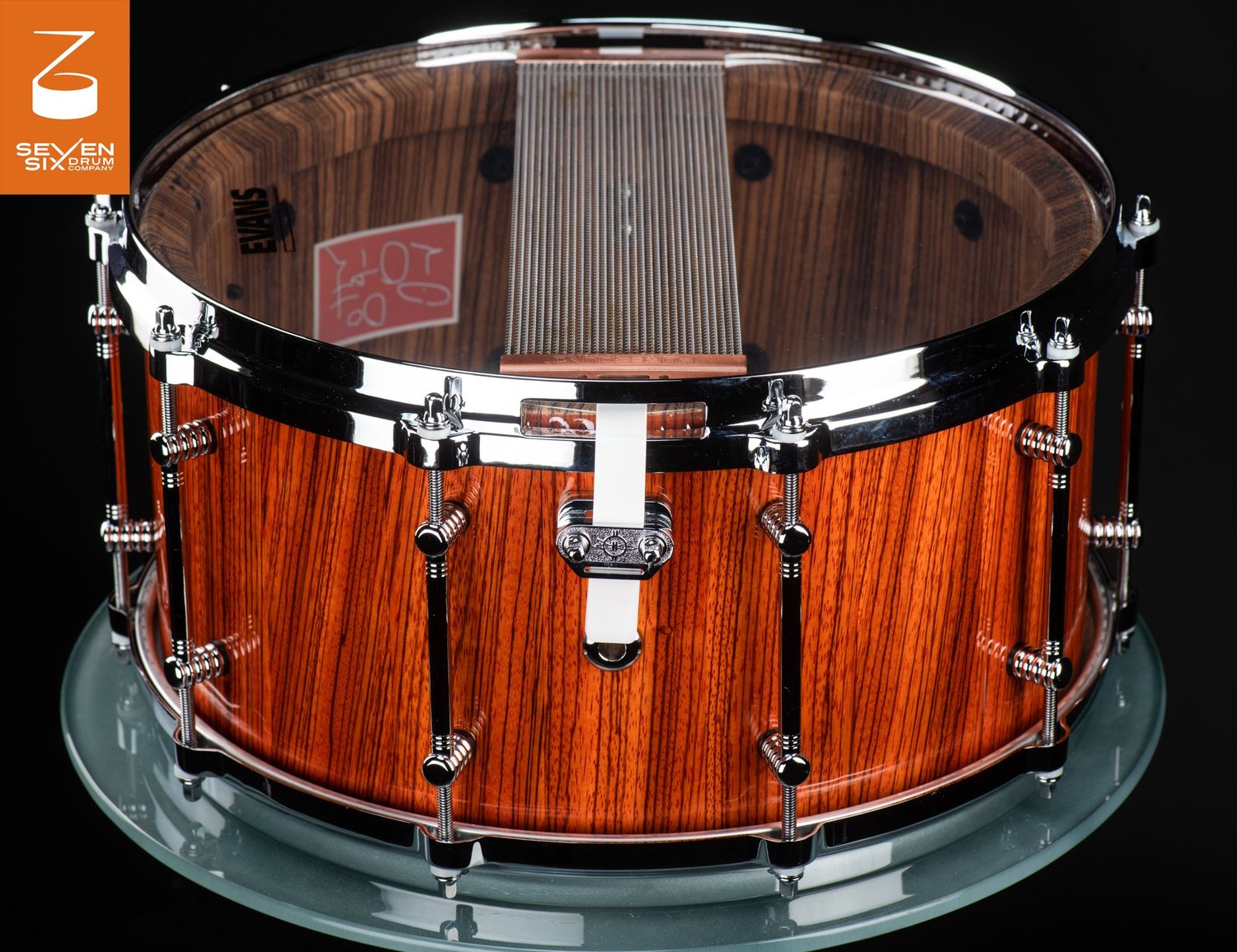 Handmade snare drum with a gloss striped wood shell, chrome tube lugs, and snare wires across a clear bottom drumhead.
