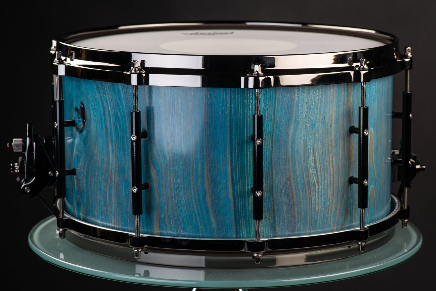 Snare drum crafted with a blue-stained vertical wood grain shell, black nickel hoops, and black tube lugs.