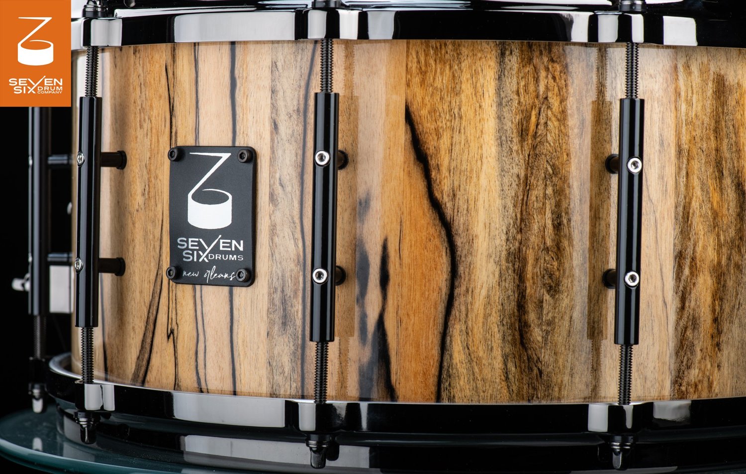 Handmade snare drum featuring a glossy natural wood shell with dark grain, sleek black tube lugs, and matching black hoops.