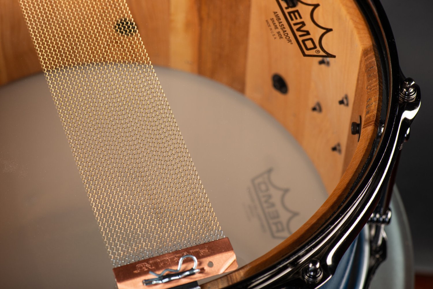 Natural finish wood snare drum featuring brass snare wires stretched across a clear drumhead and black nickel hardware.