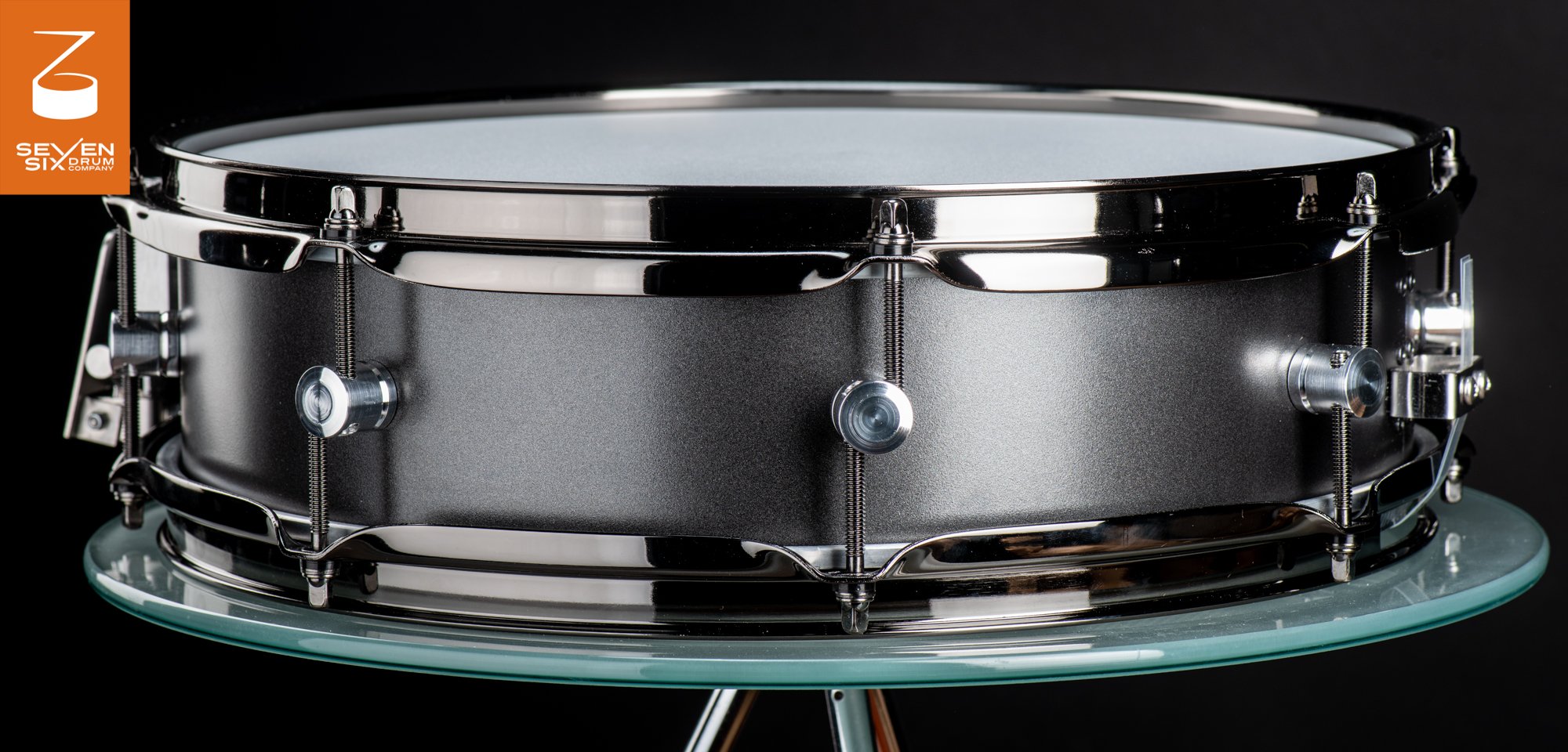 Handmade snare drum with a matte charcoal metal shell, polished black nickel hoops, and circular silver lugs.