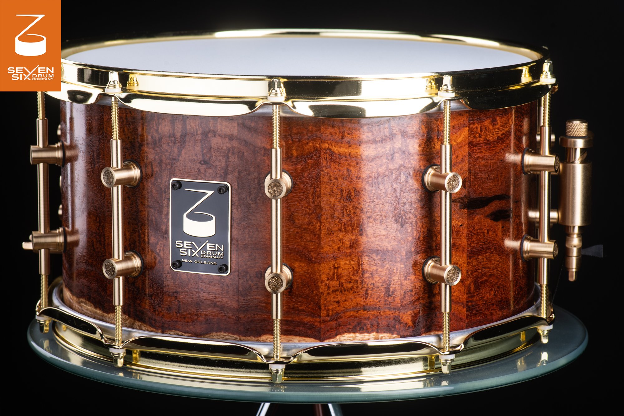 Handmade snare drum featuring a glossy figured wood shell with polished brass hoops and tube lugs.
