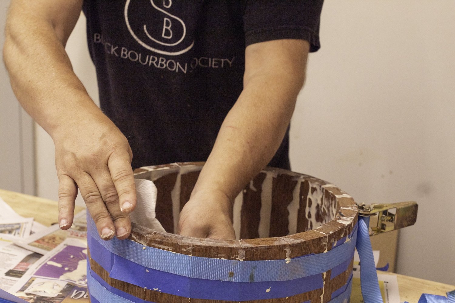 A craftsman wipes glue from an unfinished stave hardwood snare shell held together by a metal ratchet strap clamp.