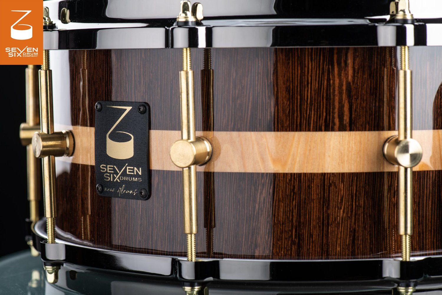 Glossy dark wood snare drum with a blonde center stripe, black hoops, and brass center-mounted lugs.
