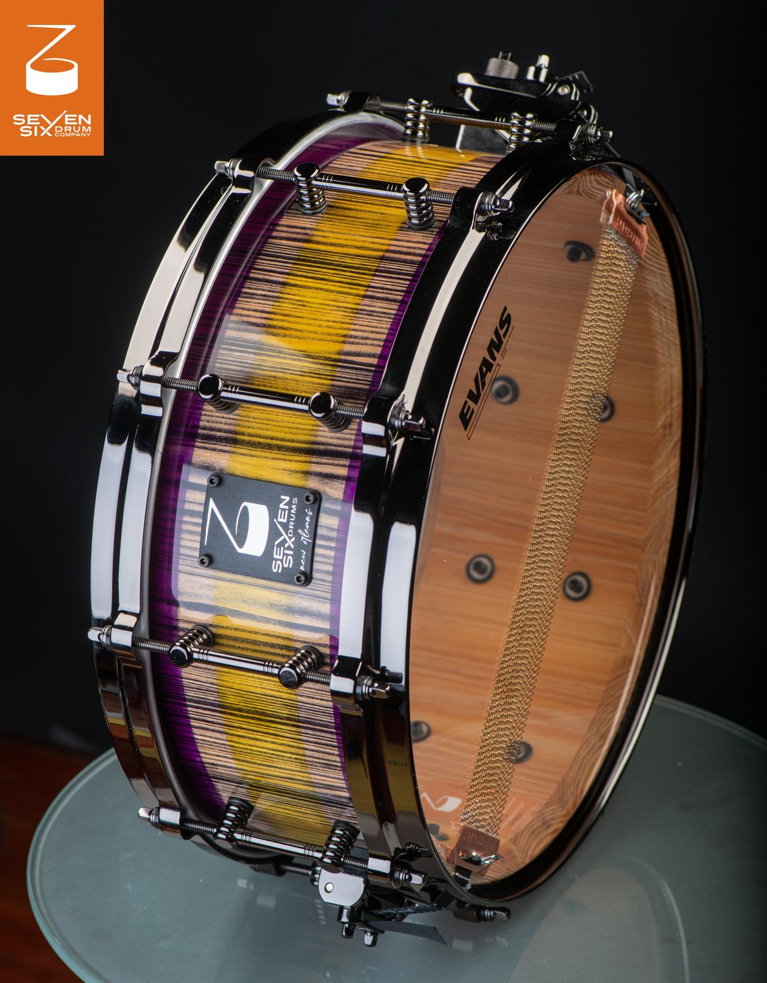 Custom snare drum featuring a glossy purple and yellow striped wood shell with black nickel rims and tube lugs.