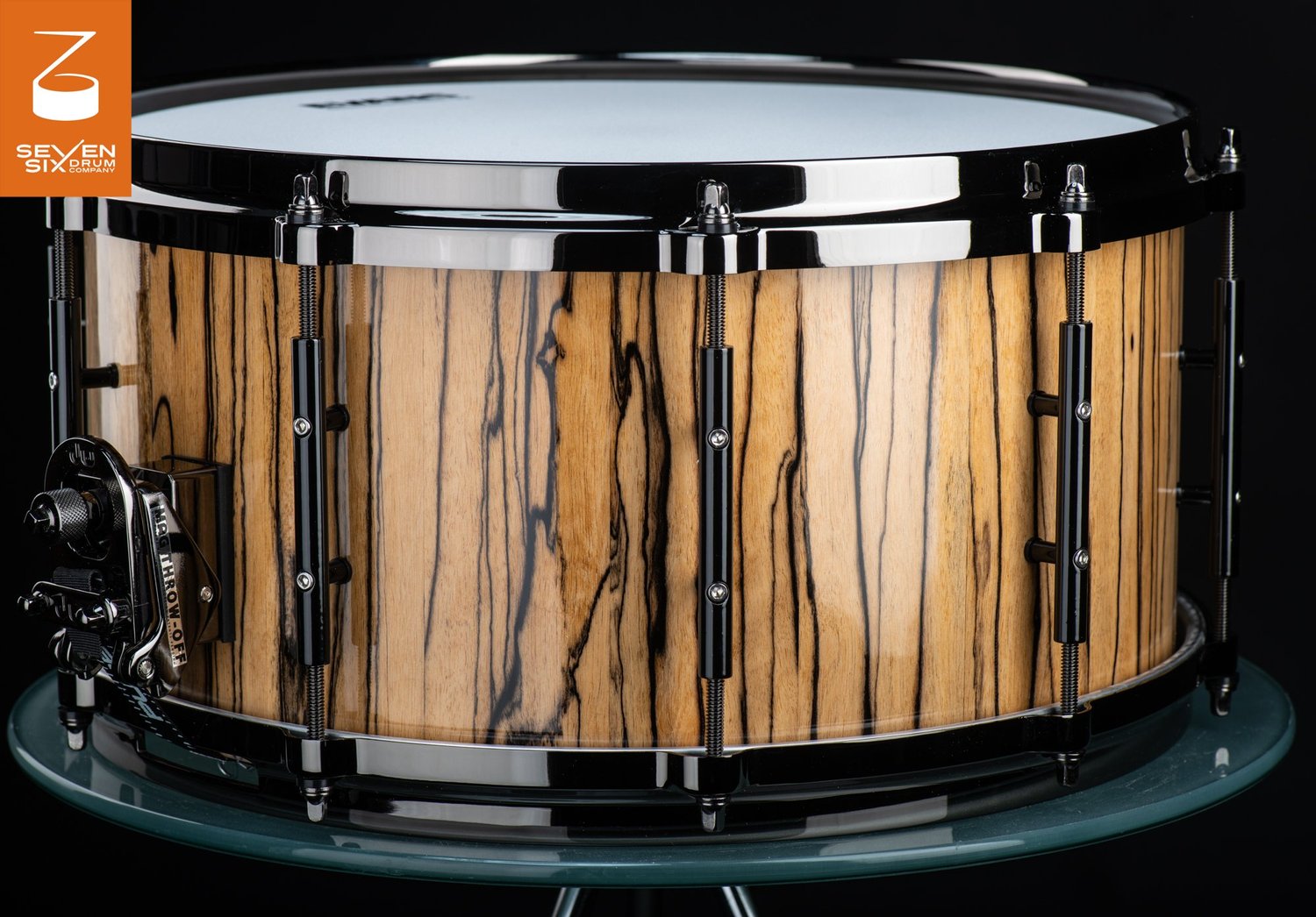 Handmade snare drum featuring a glossy striped wood shell, black nickel hoops, tube lugs, and a MAG throw-off.