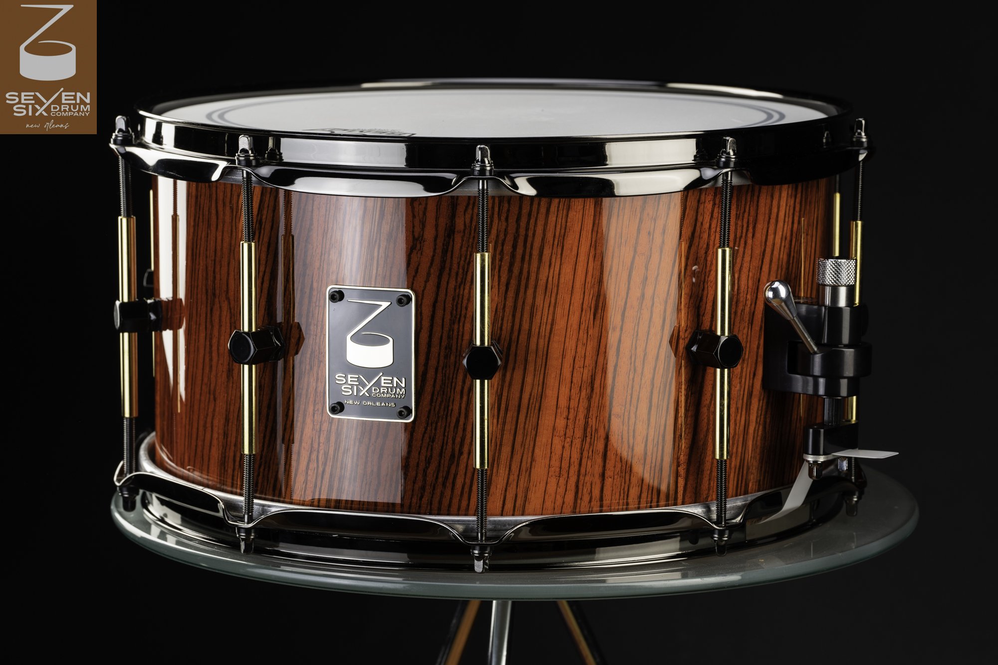 Handmade snare drum featuring a glossy reddish wood grain shell, brass tube lugs, and polished dark chrome hardware.