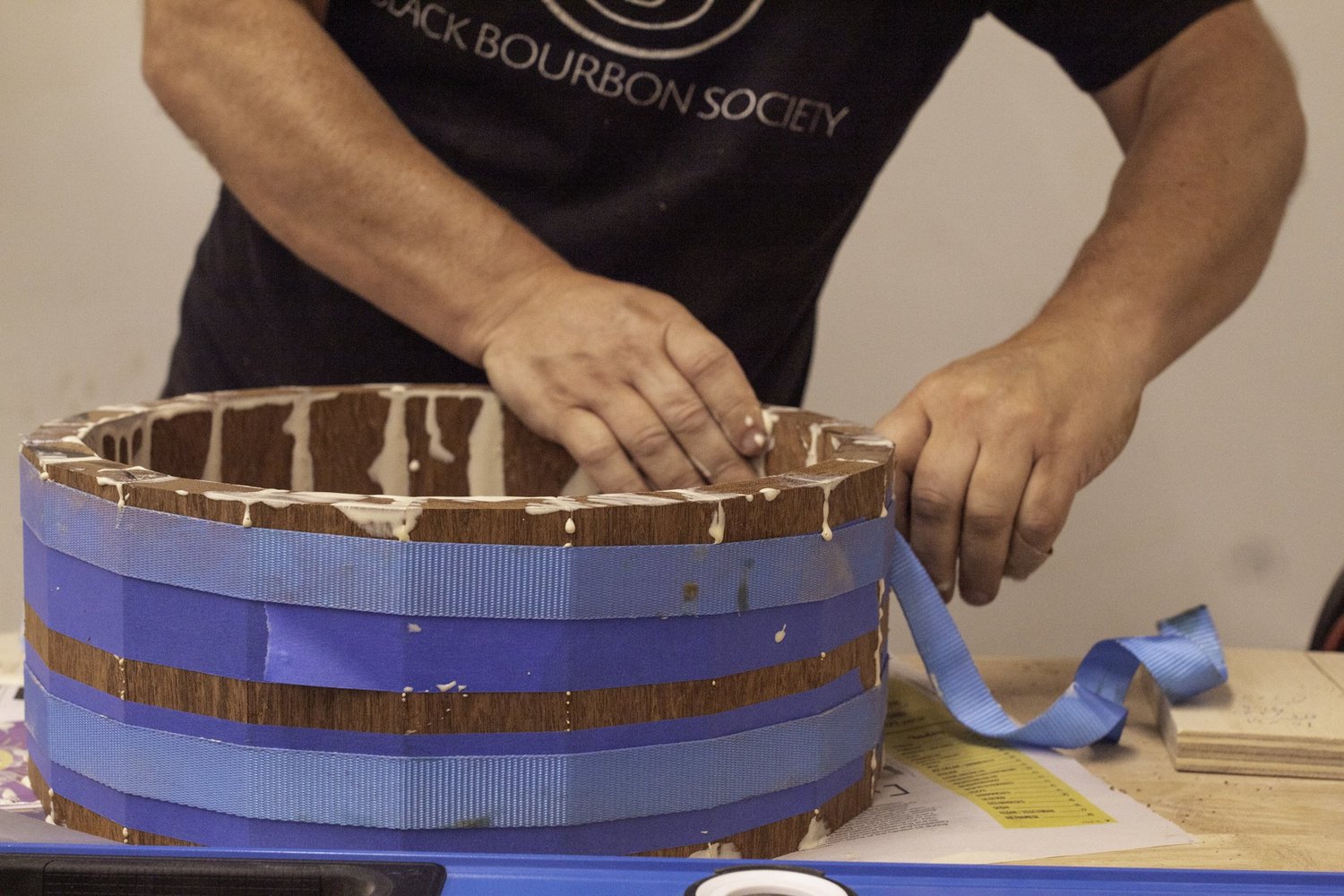 A drum maker secures an unfinished stave wood snare drum shell using blue nylon strap clamps.