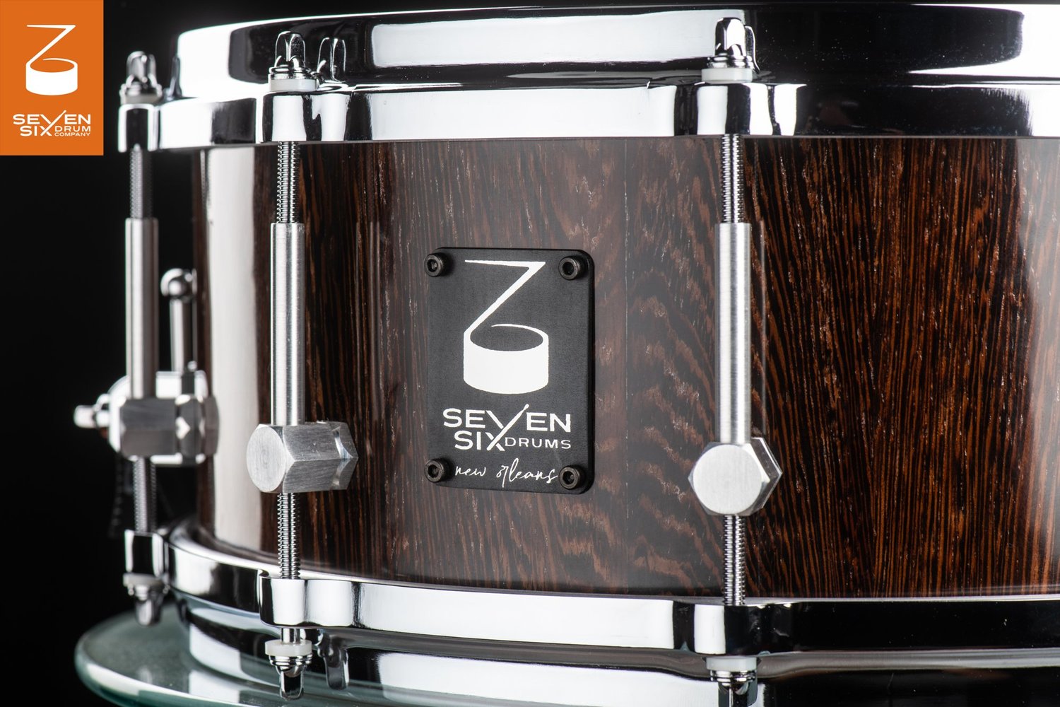 Handmade snare drum featuring a dark brown wood shell with a gloss finish, chrome hoops, hex lugs, and a black logo badge.