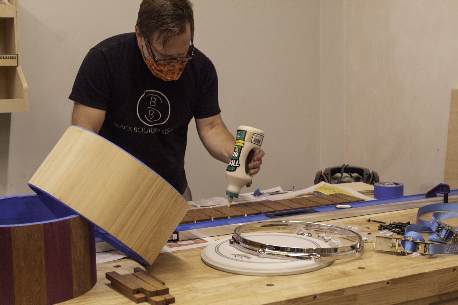 A craftsman glues natural wood staves beside unfinished drum shells and a chrome snare hoop.