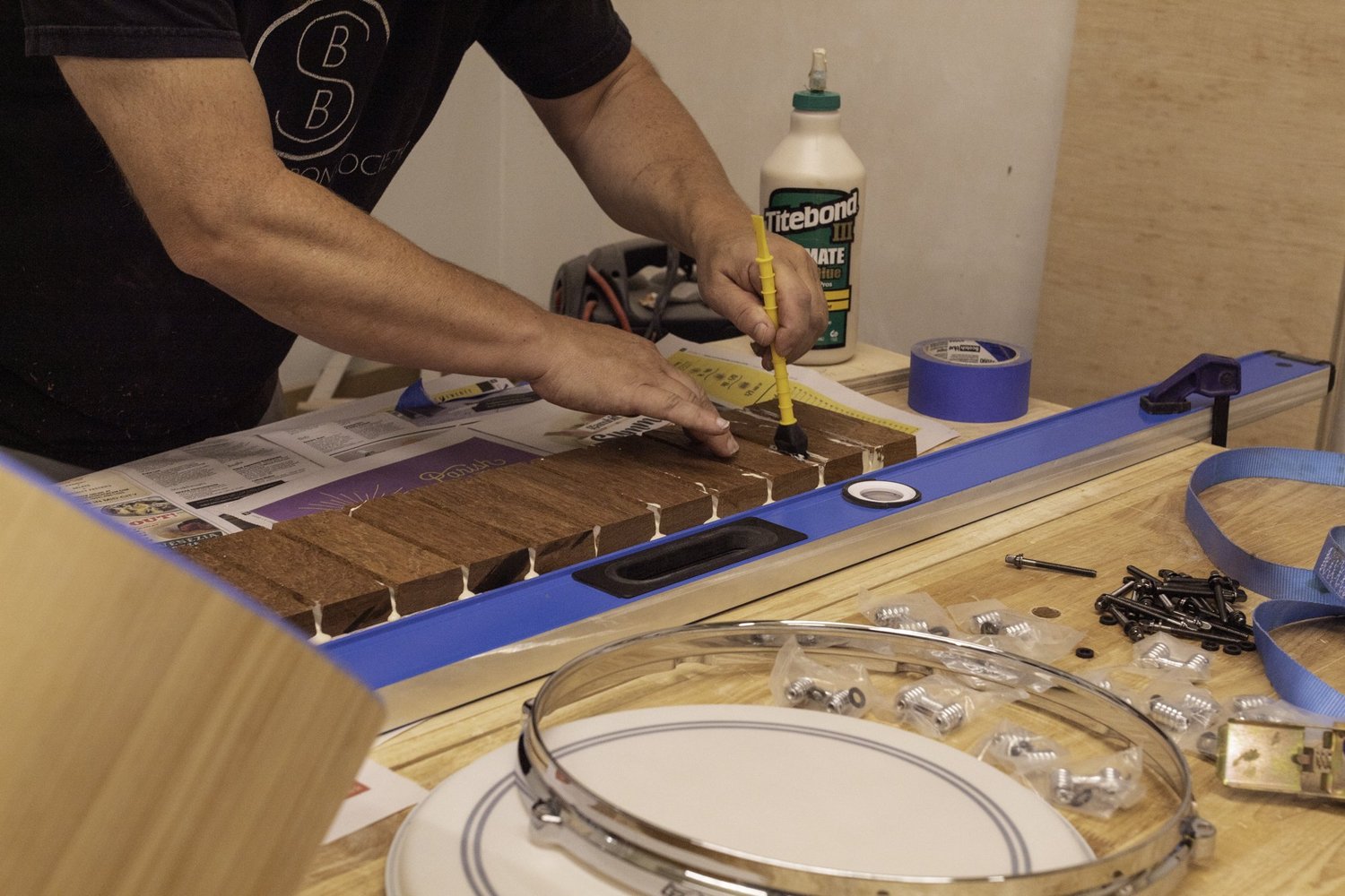 A drum maker glues raw hardwood staves on a bench beside a chrome metal hoop, lugs, and tension rods.