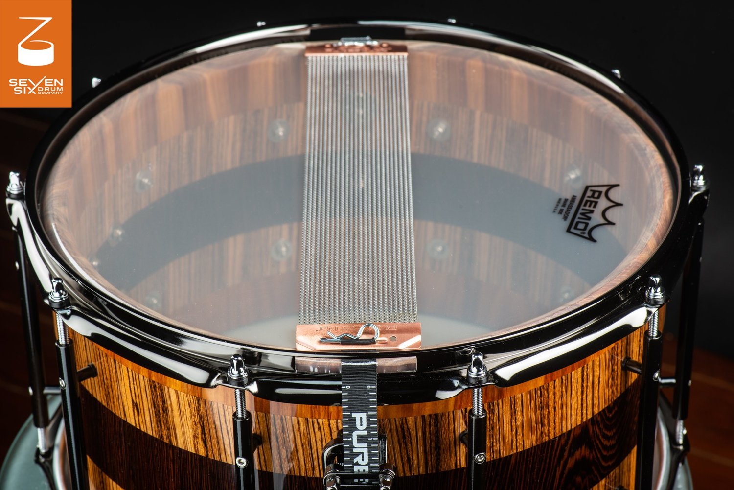 Bottom view of a glossy striped wood snare drum with black hoops, tube lugs, and wire snares across a clear drumhead.