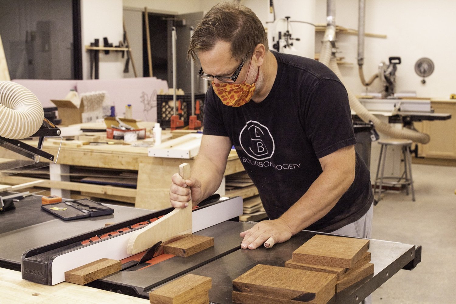 A craftsman cuts raw reddish hardwood blocks on a table saw using a push stick in a drum workshop.