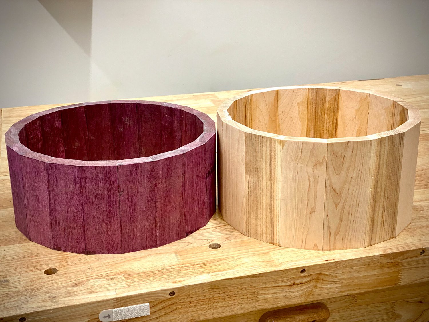 Two raw stave snare drum shells in purpleheart and light wood rest on a workbench with no hardware installed.