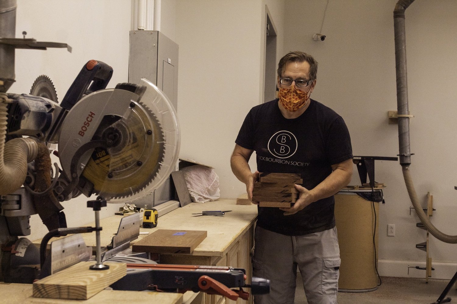 A drum maker holds cut, unfinished hardwood staves next to a Bosch miter saw in a workshop.