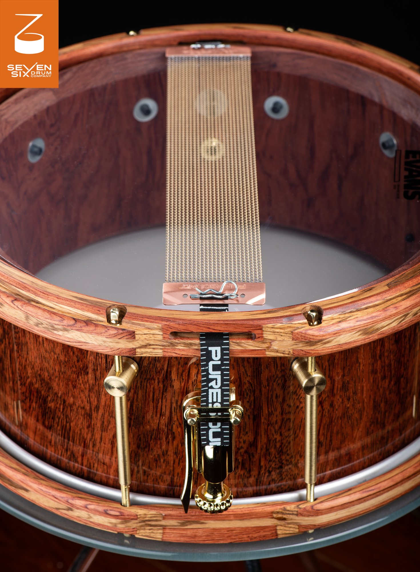 Glossy hardwood snare drum with striped wooden hoops, brass tension rods, and Puresound wire snares across a clear head.