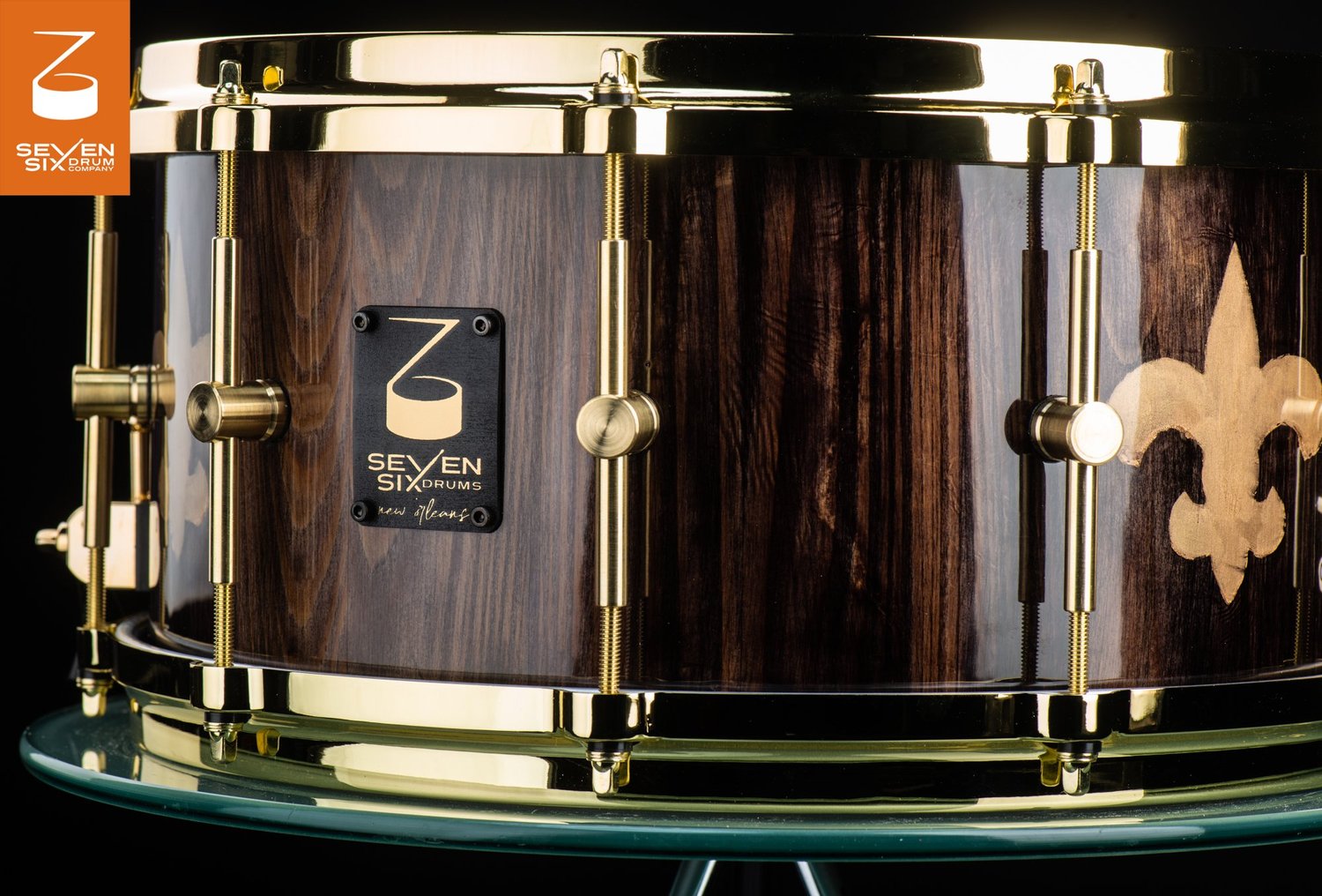 Snare drum with a glossy dark wood shell, gold fleur-de-lis inlay, and polished brass hoops and lugs.