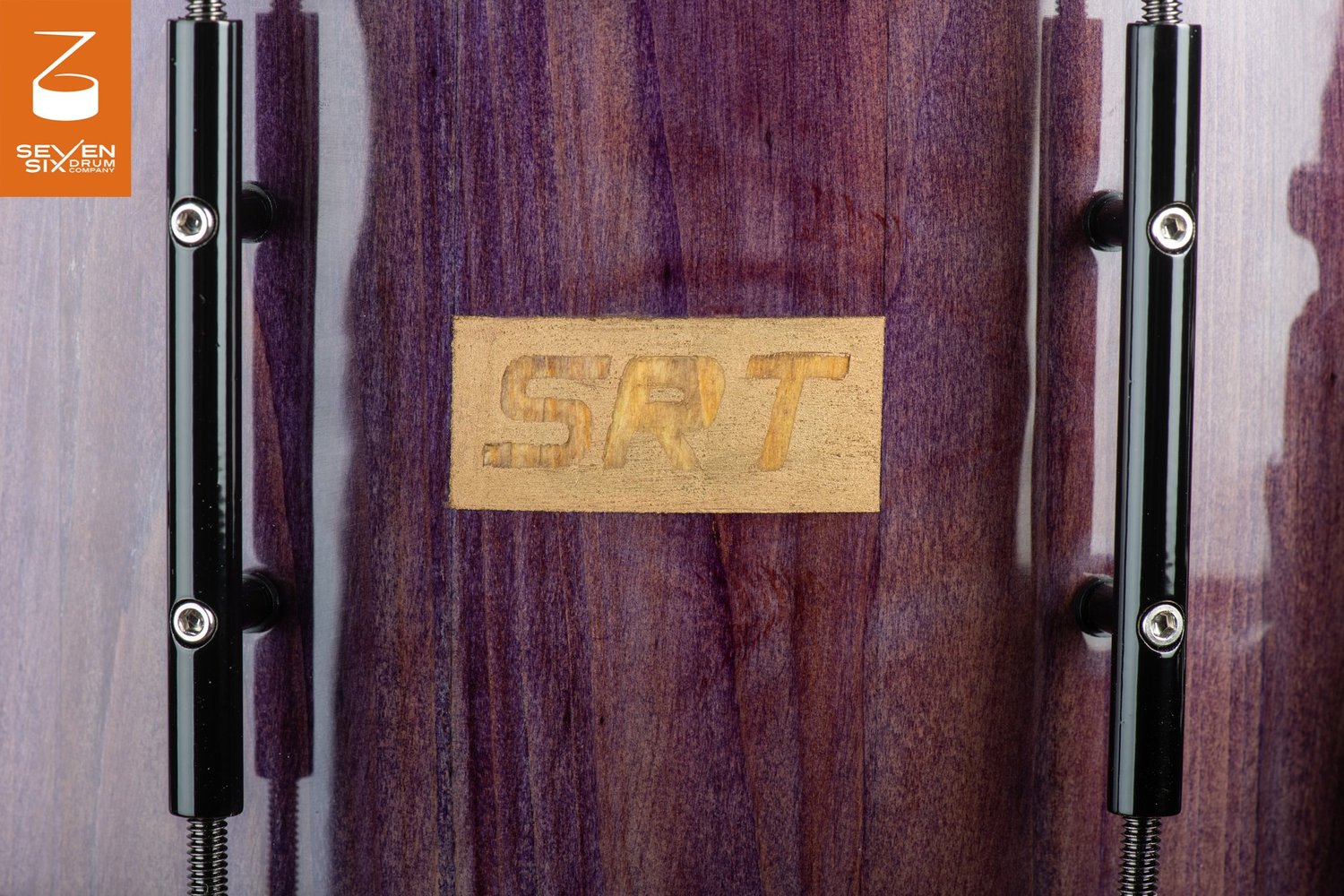 Glossy purple wood snare drum shell featuring a carved wooden logo badge flanked by black metal tube lugs.