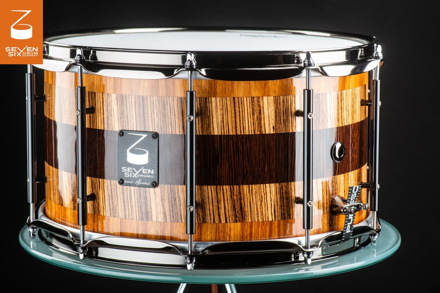 Gloss-finished wood snare drum with a dark center stripe, chrome hoops, and black tube lugs.